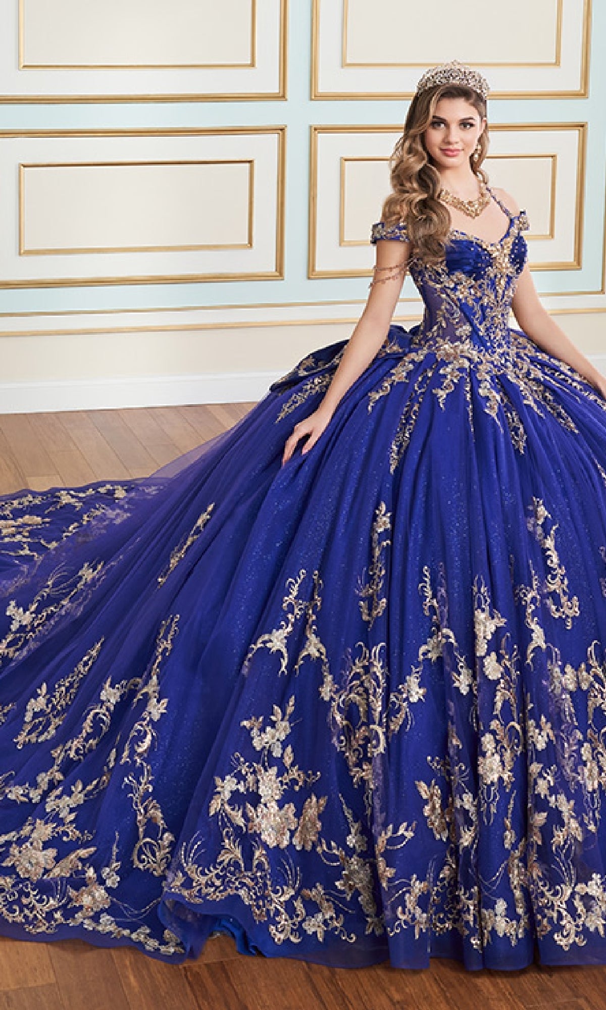 A woman in the Quinceanera Dress PR30176 By Princesa, featuring royal blue fabric with gold embroidery and metallic beading, plus a jeweled tiara, stands on a wooden floor with ornate indoor wall paneling.